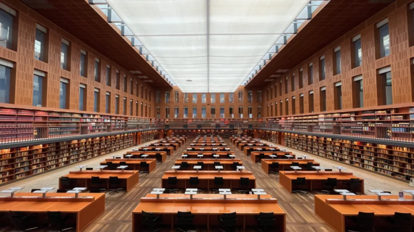 Blick in den zentralen Lesesaal der SLUB Dresden mit drei langen Tischreihen, umlaufenden Bücherregalen auf zwei Ebenen und einer lichtdurchlässigen Dachkonstruktion.